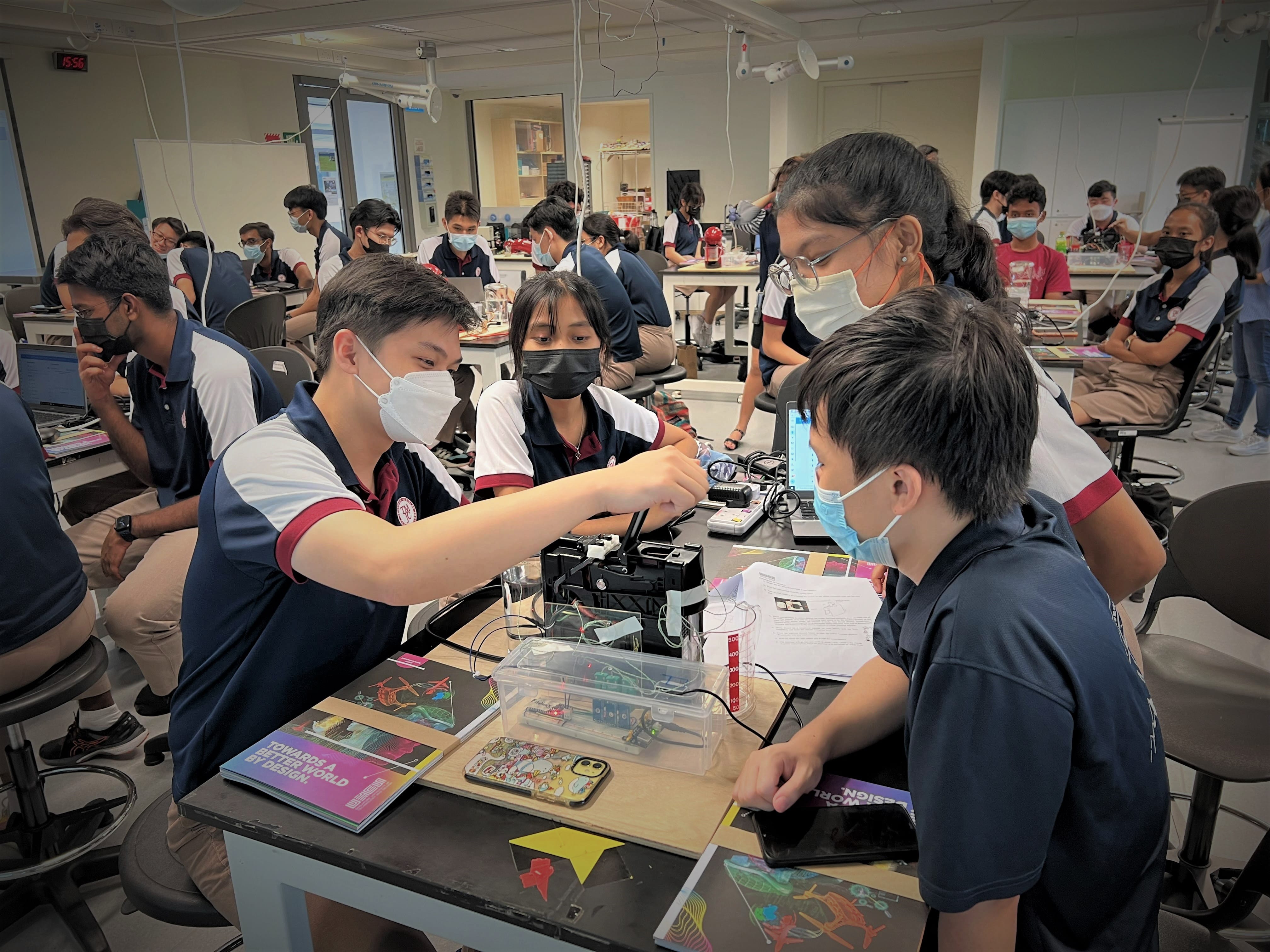 Students wearing face masks work together on an electronic device at a lab table in a busy classroom.